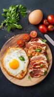 Delicious breakfast spread featuring fried egg, crispy bacon, fresh tomatoes, and herbs on a wooden cutting board photo