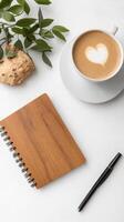 A cozy workspace featuring a cup of coffee with a heart design, an open notebook, and greenery on a clean white table photo