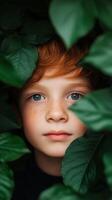 A young boy with red hair gazes curiously through vibrant green leaves in a lush garden during a sunny afternoon photo