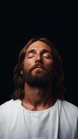 Man Jesus Christ with long hair and beard closes eyes in contemplative moment, illuminated against dark background, conveying peace and serenity photo