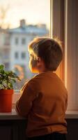 A young boy gazes thoughtfully out the window at sunset while standing beside a potted plant in a cozy indoor setting photo