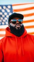 A man wearing sunglasses and a red hoodie stands confidently in front of an American flag o photo