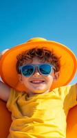A cheerful boy wearing sunglasses and a bright yellow hat enjoys a sunny day at the beach, smiling for the camera in clear blue skies photo