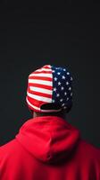 A person wearing a red hoodie and a cap featuring the American flag stands in front of a dark background during a casual indoor setting photo