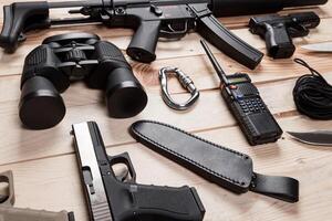 Assault rifle, gun, knife with sheath, compass and notebook with pen on table. photo