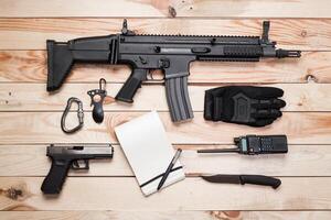 Assault rifle, gun, knife with sheath, compass and notebook with pen on table. photo