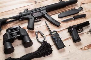 Assault rifle, gun, knife with sheath, compass and notebook with pen on table. photo