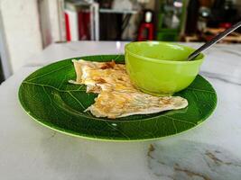 A close up of a traditional Malaysian roti canai served on a green leaf patterned plate with a green bowl and spoon. The setting appears to be a casual dining environment photo