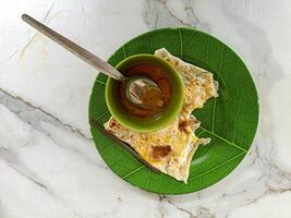 A close up of a traditional Malaysian roti canai served on a green leaf patterned plate with a small bowl of curry sauce and a spoon. The vibrant colors and textures make it visually appealing photo