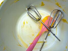 A close up of a mixing bowl with yellow batter remnants, a pink spatula, and two metal beaters. This image captures the aftermath of baking, highlighting the tools and traces of the process photo