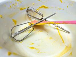 A close up of dirty kitchen utensils, including beaters and a spatula, covered in yellow batter in a mixing bowl. The image highlights the aftermath of baking photo