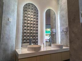 Modern bathroom interior with two round sinks, sleek faucets, and illuminated mirrors. The design features a textured wall and a unique geometric pattern, creating a stylish and contemporary look photo
