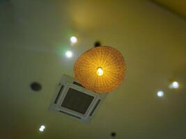 A warm, woven rattan lampshade with a glowing bulb hangs from a ceiling, surrounded by recessed lights and an air conditioning vent, creating a cozy ambiance photo