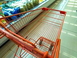 An empty red shopping cart in a grocery store aisle, with shelves of packaged goods in the background. The cart is positioned near the refrigerated section, typical shopping experience photo