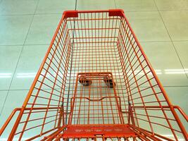 An empty orange shopping cart with a wireframe design is seen from above on a tiled floor. The cart is positioned in a well-lit, clean environment, likely a supermarket or store photo