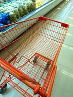 An empty orange shopping cart in a grocery store aisle, with shelves of packaged food items in the background. The cart is positioned near a refrigerated section photo