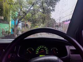 A view from inside a car during a rainy day, showing the wet windshield with raindrops, the steering wheel, and the dashboard with illuminated gauges photo