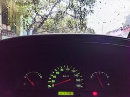 A car dashboard showing speedometer, fuel gauge, and temperature gauge with raindrops on the windshield, indicating a rainy day photo