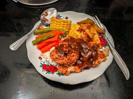 A plate of fried chicken with gravy, rice, corn, green beans, carrots, and fried potatoes on a floral patterned plate, accompanied by a spoon and fork on a black surface photo