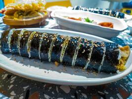 A close up of a sliced Korean gimbap roll on a white plate, garnished with sesame seeds. The background shows other dishes, highlighting a traditional Korean meal setting photo