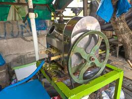 A rustic sugarcane juicer with a large green wheel, set outdoors. Surrounding items and a tree create an authentic countryside scene photo
