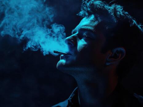 Man exhales smoke in a dimly lit setting with deep blue lighting during an evening moment of reflection photo