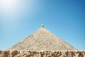 Red sphere balanced atop a pyramid under a clear blue sky with faded colors reflecting a serene atmosphere photo