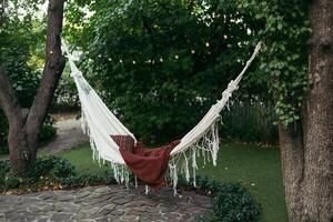 Tranquil backyard with a cozy hammock tied between trees, featuring soft blankets, surrounded by greenery and string lights. photo