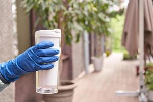 Hand in blue glove holding reusable cup on sidewalk with potted plants and umbrella in background. photo
