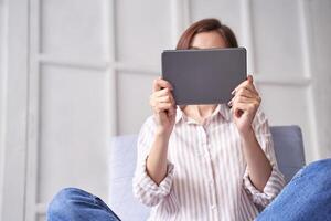 Young asian female sitting indoors using tablet for browsing or reading in casual home setting. photo