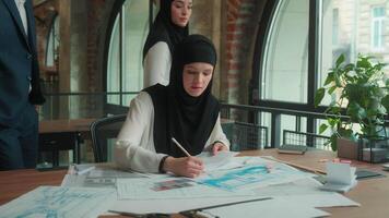 Muslim designer architect in hijab working in office drawing blueprints architecture project on desk business man women multiethnic team colleagues engineers help cooperate discuss building plan draft video