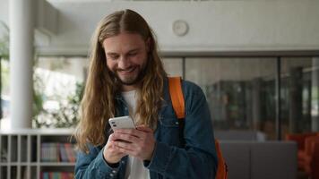 Male indoors in academy university laugh having fun funny reaction holding mobile phone watching SMS using mobile phone Caucasian man guy student smiling laughing joy humorous social media app video