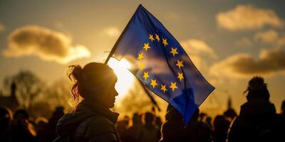 A silhouette of a woman holding a European Union flag during sunset with soft clouds and a crowd in the background photo