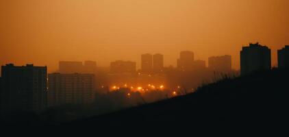 City lights glowing through layers of pollution create moody atmosphere at dusk. silhouettes of buildings stand against hazy orange sky, evoking sense of urban mystery photo