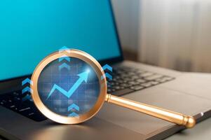 Growth and success arrow on a magnifying glass near a laptop. Growth of indicators, optimistic forecasts. Stock prices, trading on the stock exchange. Economy photo