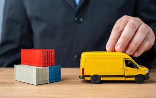 A businessman pushes a delivery van past a stack of shipping containers. The concept of delivery and transportation of goods and parcels to the end customer. Logistics and speed. photo