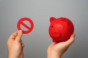 Red piggy bank and prohibitory sign no in hands on gray background. Overheated financial market. Difficult economic conditions. Heavy burden on budget. Financial restrictions and tighter regulation. photo
