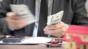 Businessman counting dollar bills at a desk with a calculator, documents, and small toy models of a house and car, symbolizing financial planning, budgeting, or property investment video