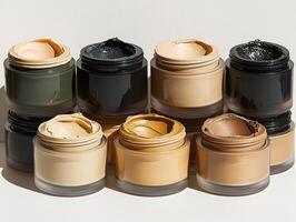 Cream eyeshadow pots in shades of champagne olive green and bronze are artfully arranged against minimalist white backdrop. creamy texture glimmers softly in light photo