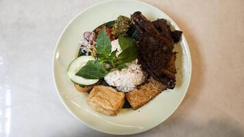 Traditional Indonesian Kecombrang Rice with Fried Spiced Duck Tempeh Tofu Assorted Sambals Fresh Basil and Cucumber Served on a Light Green Plate on Neutral Table Top View. photo