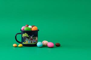 Colorful Easter chocolate eggs in enamel mug against green background. Minimal Easter concept. Creative festive spring holiday layout. Easter aesthetic. photo