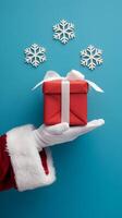 santas mano con regalo caja, copos de nieve flotar, alegre fiesta escena en azul antecedentes vertical móvil fondo de pantalla foto