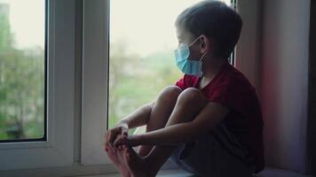 Sad boy in medical mask sits on windowsill and looks out window, self quarantine 02 video
