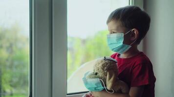 Boy and his teddy bear both in protective medical masks sits on windowsill and looks out window 01 video