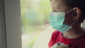 Boy and his teddy bear both in protective medical masks sits on windowsill and looks out window 02 video
