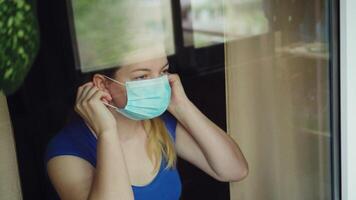 Woman in a medical face mask looks out of the window and speak at mobile 02 video