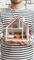 Man holds 3D house model, architectural blueprint backdrop highlights design focus Vertical Mobile Wallpaper photo