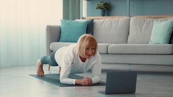 A senior woman is engaging in a workout routine at home, demonstrating a plank position. She focuses on her health while following an online fitness session in a bright room. video