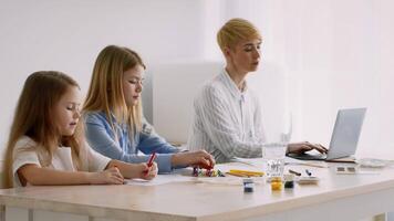 Two children are focused on drawing and crafting with colorful materials, while an adult is working on a laptop at a sunny table. The atmosphere is filled with creativity and concentration. video