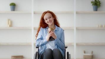 A cheerful redhead girl with a disability is seated in her wheelchair, smiling happily while holding a trophy. The setting is a well-lit room with minimal decor, suggesting a cozy atmosphere. video